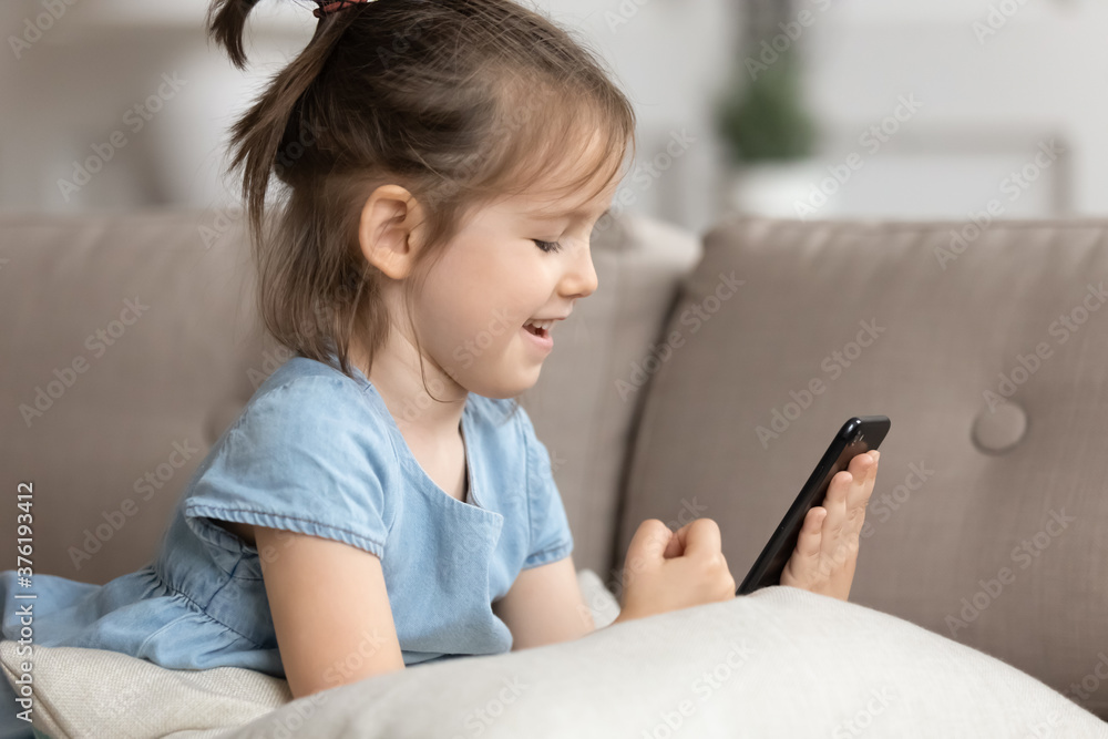Close up cute little girl lies on sofa holds smart phone make videocall spend time on internet. New generation addicted with modern tech problem, gadget dependence, parental control system app concept Close up cute little girl lies on sofa holds smart phone make videocall spend time on internet. New generation addicted with modern tech problem, gadget dependence, parental control system app concept
