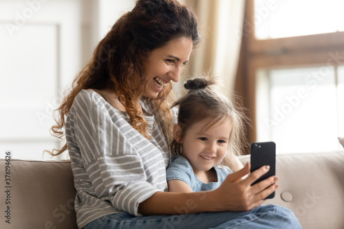 Mum and curious little daughter sit on couch hold cell phone watch cartoons use new funny app, during video call conference mom and kid girl laughing enjoy distant talk, parental control, secure usage