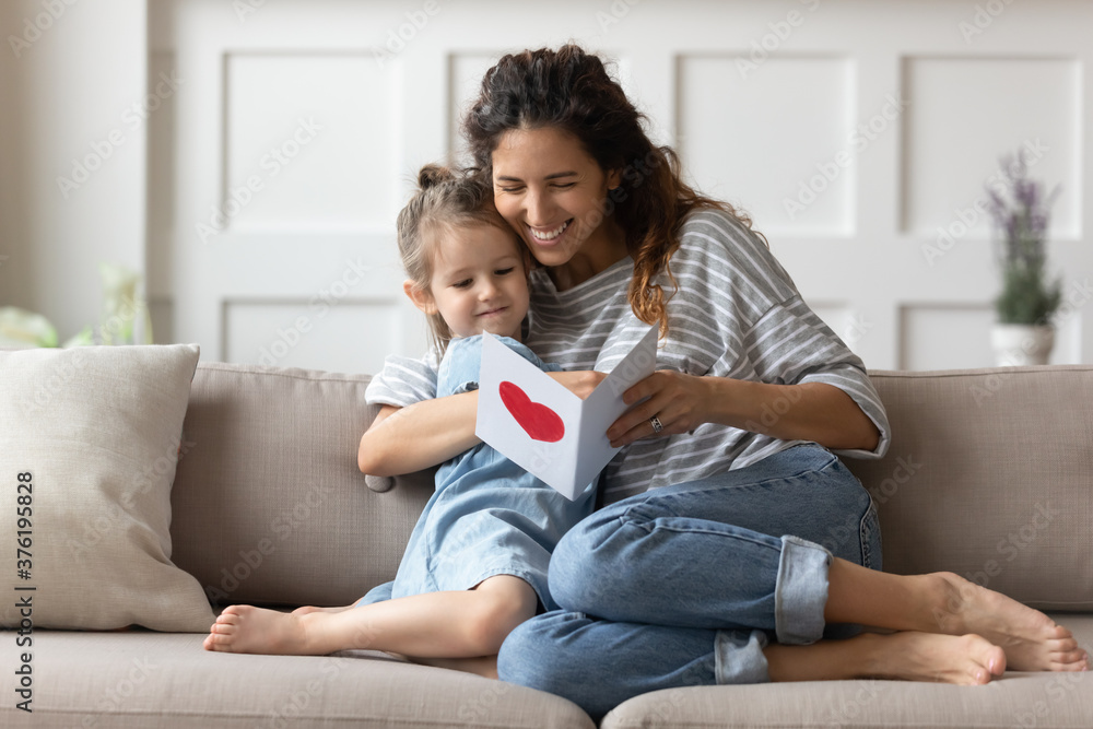 Mum and little daughter sit on sofa in cozy living room hold paper card read wishes on handmade postcard. Attentive small kid made gift on International Women Day to loving mom, Mother Day celebration Mum and little daughter sit on sofa in cozy living room hold paper card read wishes on handmade postcard. Attentive small kid made gift on International Women Day to loving mom, Mother Day celebration