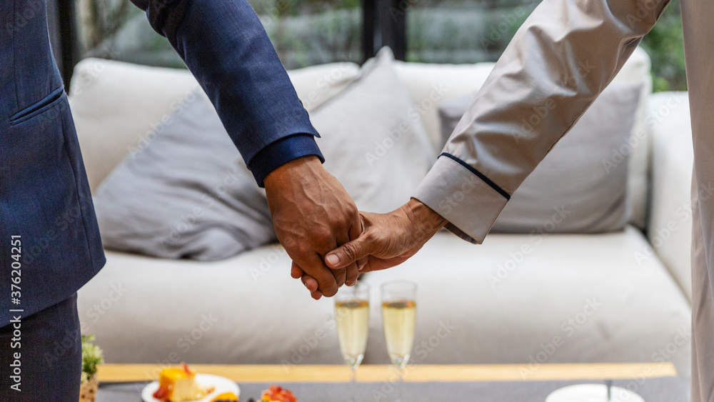 Two gays from different cultures holding hands together while celebrating partnership in a living corner at home. Two men holding hands to support each other over drinking table Two gays from different cultures holding hands together while celebrating partnership in a living corner at home. Two men holding hands to support each other over drinking table