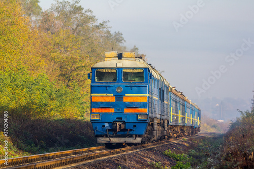 An old diesel locomotive with a commuter train arrives at a stopping point. Sunny autumn day.