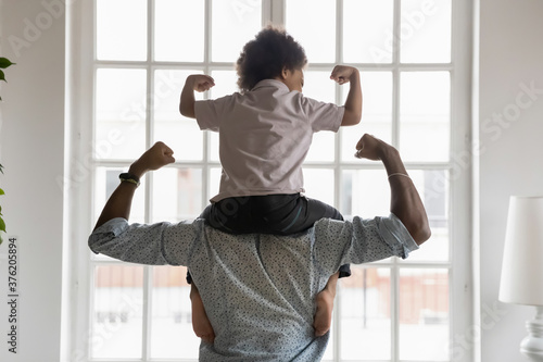 Murais de parede Back side view young african american father holding on shoulders little preschool funny kid son, showing biceps