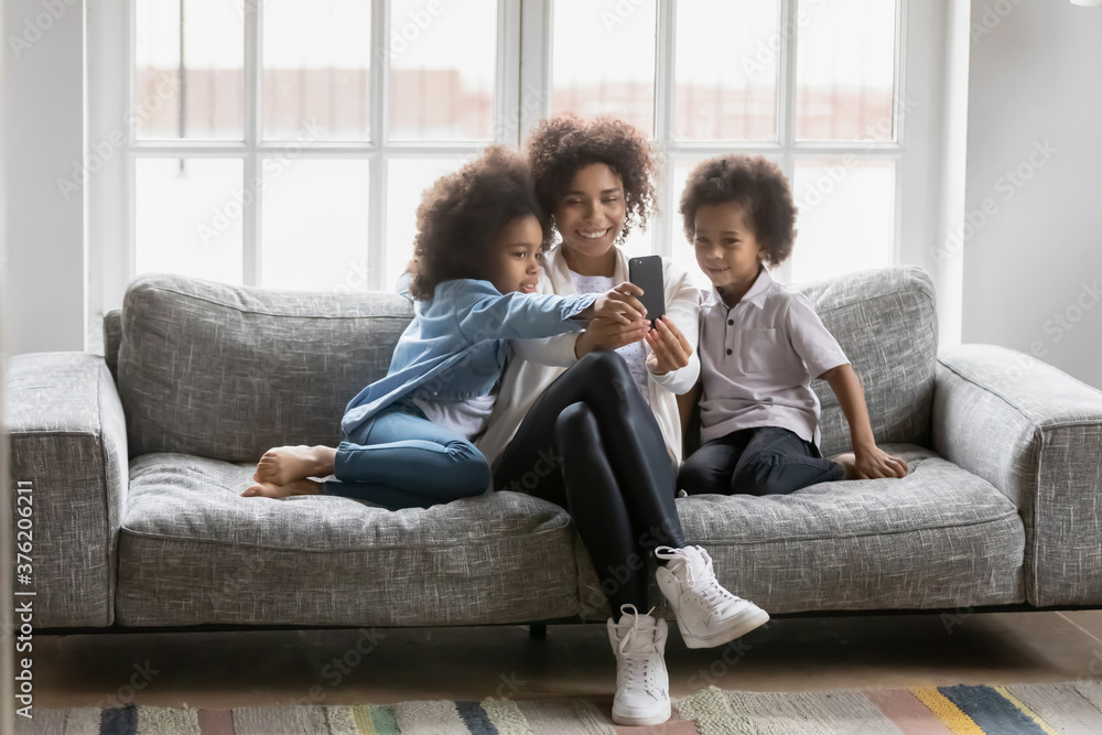 Full length addicted to technology smiling beautiful african american woman showing funny mobile application to happy small daughter ands preschool son, relaxing together on comfortable couch at home.