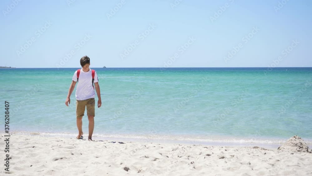 eenager on the background of the azure sea walks along the beach on a Sunny summer day. He's wearing a t-shirt and shorts. eenager on the background of the azure sea walks along the beach on a Sunny summer day. He's wearing a t-shirt and shorts.