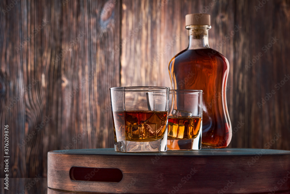 Two square glasses of iced whiskey and a full bottle of whiskey stand on a tray against an old wooden wall. Two square glasses of iced whiskey and a full bottle of whiskey stand on a tray against an old wooden wall.