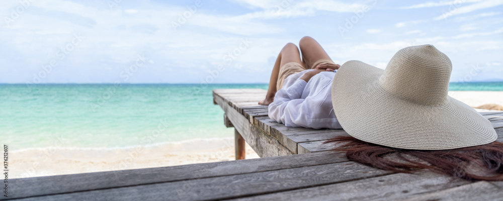 Asian woman with straw hat relaxing on wooden terrace at tropical beach, sea view, Summer Beach Vacation Concept.