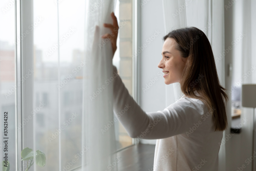 Side view happy attractive woman opening curtains, admiring cityscape at home or hotel room. Smiling millennial girl enjoying morning leisure weekend time or new day start, planning future indoors. Side view happy attractive woman opening curtains, admiring cityscape at home or hotel room. Smiling millennial girl enjoying morning leisure weekend time or new day start, planning future indoors.