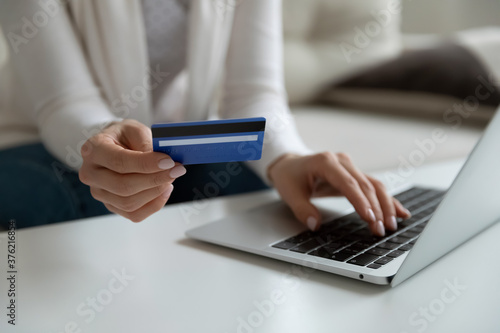 Close up young woman holding plastic banking credit debit card, involved in online shopping on computer at home. Female client entering payment information cvv code confirming purchase indoors.