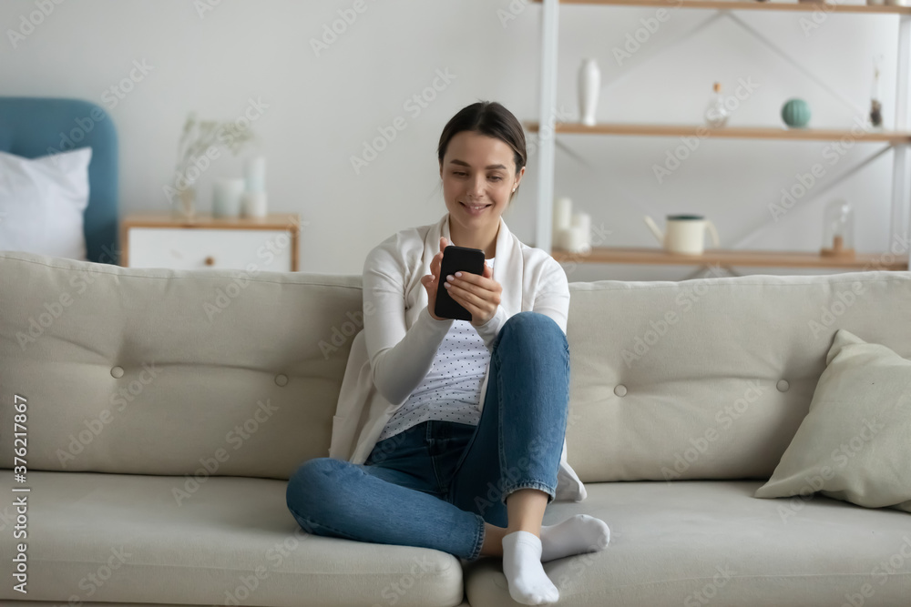 Full length beautiful young woman sitting on comfortable couch, using entertainment applications on smart phone. Happy millennial girl looking at screen, communicating in social networks with friends.