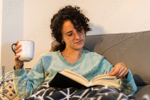 Stock photo of a beautiful and happy young woman with curly hair and glasses reading a book at home, sitting on the coach. Curly girl with soft blanket and winter pajama reading on a winter afternoon. photo