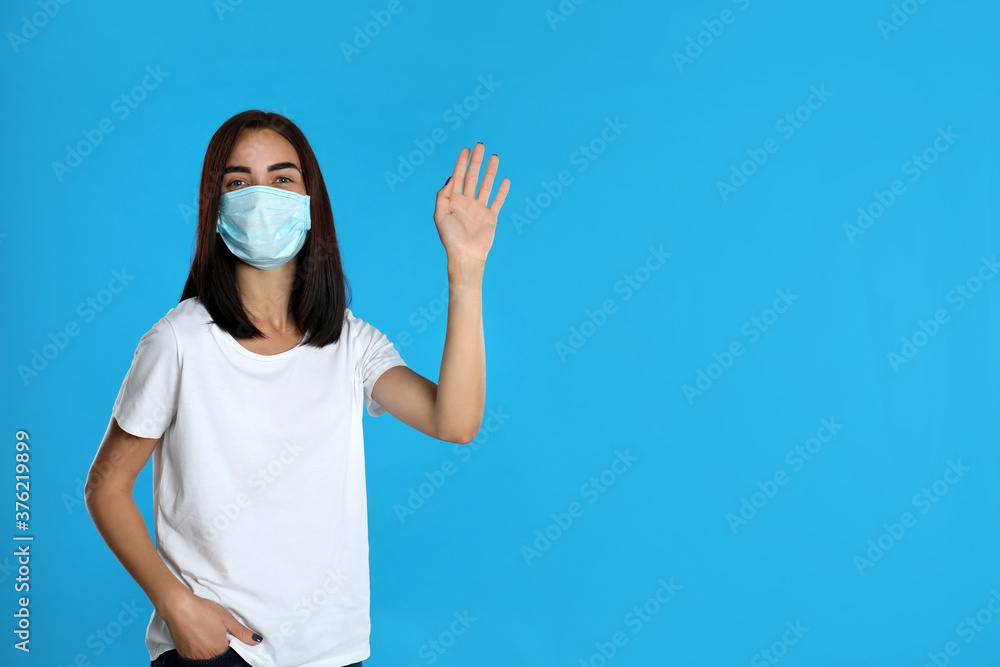Young woman in protective mask showing hello gesture on light blue background, space for text. Keeping social distance during coronavirus pandemic Young woman in protective mask showing hello gesture on light blue background, space for text. Keeping social distance during coronavirus pandemic
