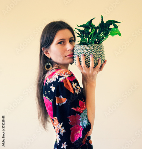 Beautiful young woman in a boho dress with kimono sleeves in a colorful floral pattern with a climbing plant with twisted leaves - Epipremnum, in a designer pot photo