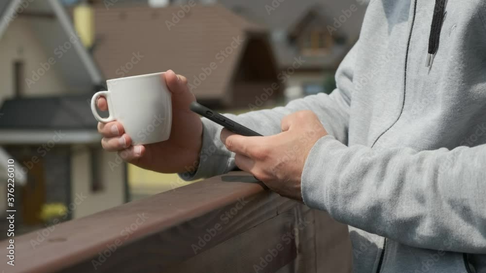 Man using his smartphone and takes a white cup in another hand on the balcony Man using his smartphone and takes a white cup in another hand on the balcony