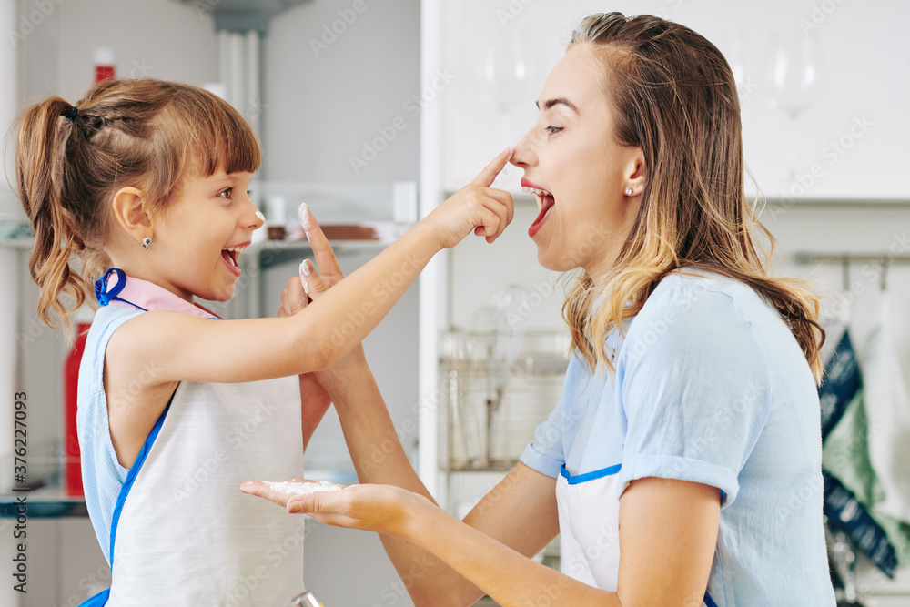 Happy pretty young woman and her little daughter playing with flour when baking pastry in kitchen