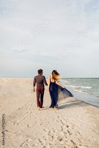 View from back. Elegant couple in love walking along the beach. Romantic moments. White sand and ocean waves. Tropical vacation. Full height.
