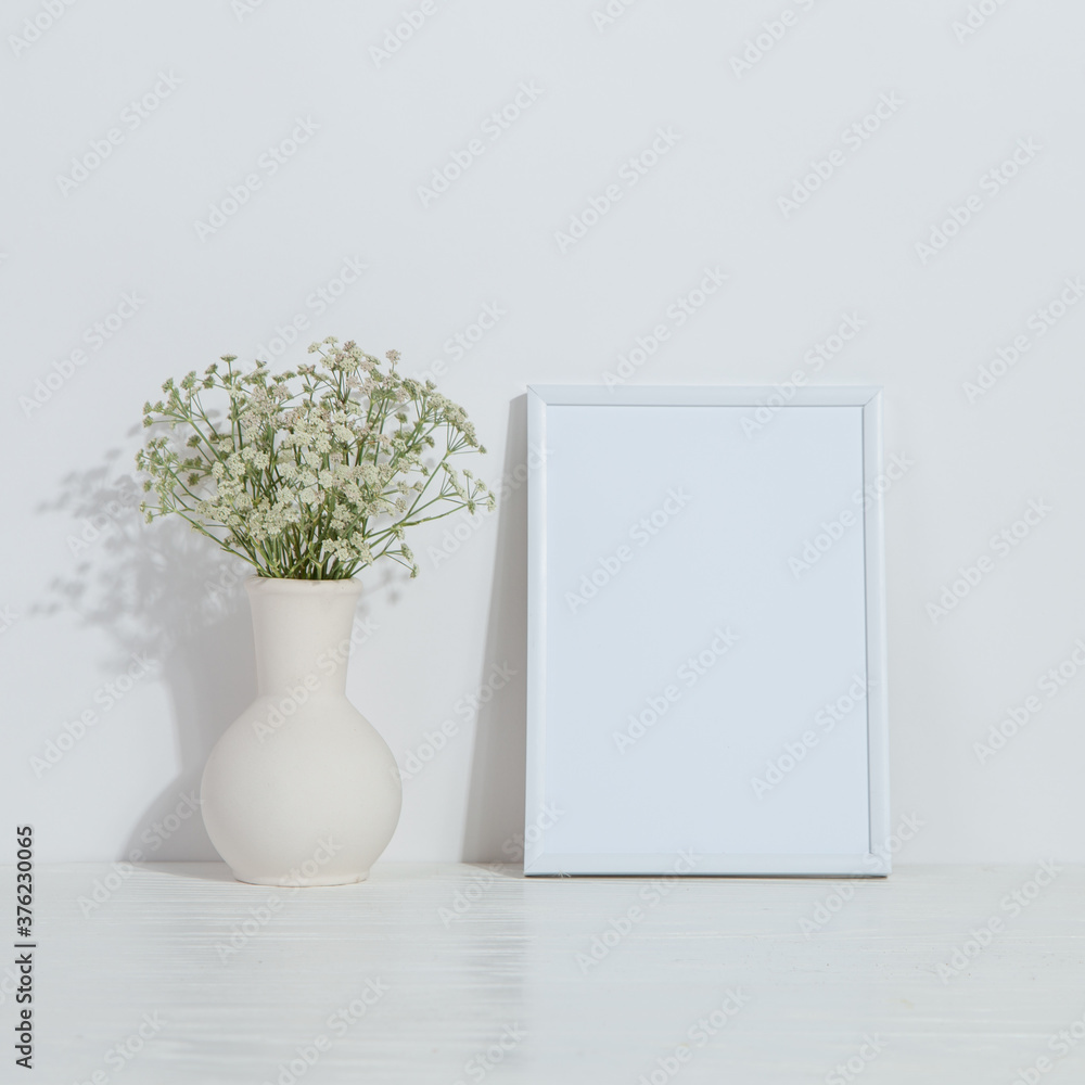A vase with flowers and a white photo frame on the table. Light stylish background. Preparation for inscriptions, copy space. A vase with flowers and a white photo frame on the table. Light stylish background. Preparation for inscriptions, copy space.