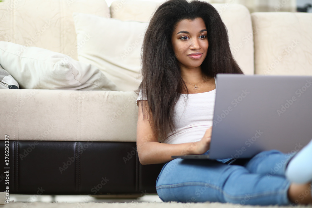 Fototapeta premium African woman sits at home next to sofa and holds laptop. Remote work around the world concept