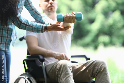 Man in wheelchair sits and holds dumbbell in his hand. The woman controls exercises. Sport for people with disabilities concept