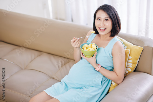Portrait of happy young Asian pregnant sitting on sofa at home and woman eating fresh pineapple slices