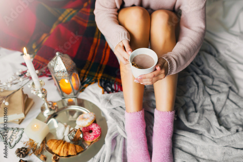 Top view image of young slim woman enjoying morning time in her bed, wearing warm cozy woolen sweater and pink socks, holding big cup of coffee.
