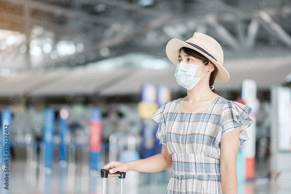 Young female wearing face mask with luggage checking flight time in airport, protection Coronavirus disease infection, Asian woman traveler ready to travel. New Normal and travel under COVID-19