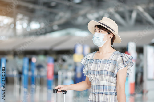 Young female wearing face mask with luggage checking flight time in airport, protection Coronavirus disease infection, Asian woman traveler ready to travel. New Normal and travel under COVID-19 © Jo Panuwat D