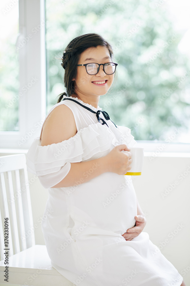 Beautiful young pregnant woman in glasses sitting on chair by window, drinking tea and smilig at camera Beautiful young pregnant woman in glasses sitting on chair by window, drinking tea and smilig at camera