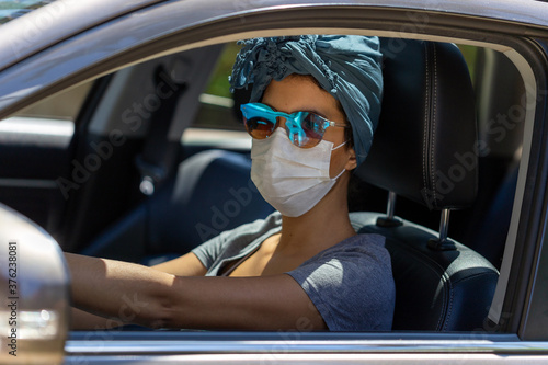 A woman driving a car wearing a mask, scarf in her hair and sunglasses to protect herself from contamination by corona virus. Pandemic. Essential services. Social isolation. Health care.  © Fernando Calmon