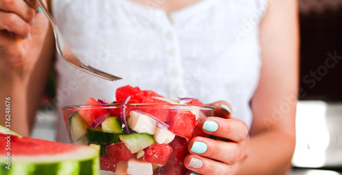 Young woman eating fresh delicious juicy dietary salad with watermelon, feta cheese, cucumber and red onions on the kitchen at home. Close-up. Side view. Healthy food concept.