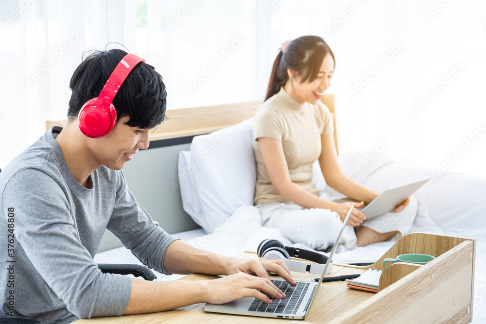 A man working with notebook wearing a bluetooth headphones to listen chatting with colleagues. His girlfriend is sitting in bed working together. They work from home. A man working with notebook wearing a bluetooth headphones to listen chatting with colleagues. His girlfriend is sitting in bed working together. They work from home.