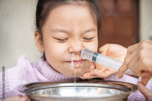 Little asian girl saline nasal wash nose at home. Asia mother making nasal wash for her kid girl by flushing nose cleaning with syringe and saline to treat the flu. health care medical people concept.