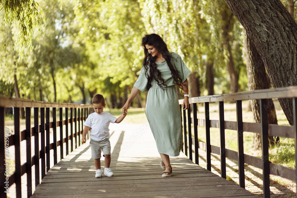 Beautiful mother with happy son walking at the park, adorable woman with joyful kid making first steps, mommy gentle looking at the boy, smiling, motherhood and childhood concept