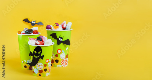 Halloween trick or treat sweets. Paper cups with colourful candies inside and paper silhouettes of bats, ghosts on bright yellow background. Spooky holiday symbols. Copy space. Wide banner.
