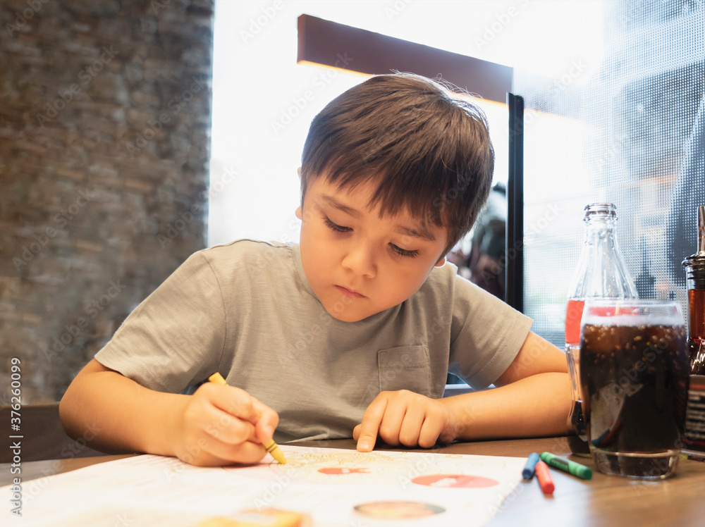 Portrait kid drawing on white paper while waiting for food inrestuarant,Selective focus of kid enjoy colouring with yellow crayon on paper, Activity for children with family Portrait kid drawing on white paper while waiting for food inrestuarant,Selective focus of kid enjoy colouring with yellow crayon on paper, Activity for children with family