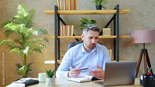 Gray-haired man in home office wears wireless earphones studying remotely online video conference call with tutor, distance education. Guy looks laptop computer talks with teacher gestures with hands photo