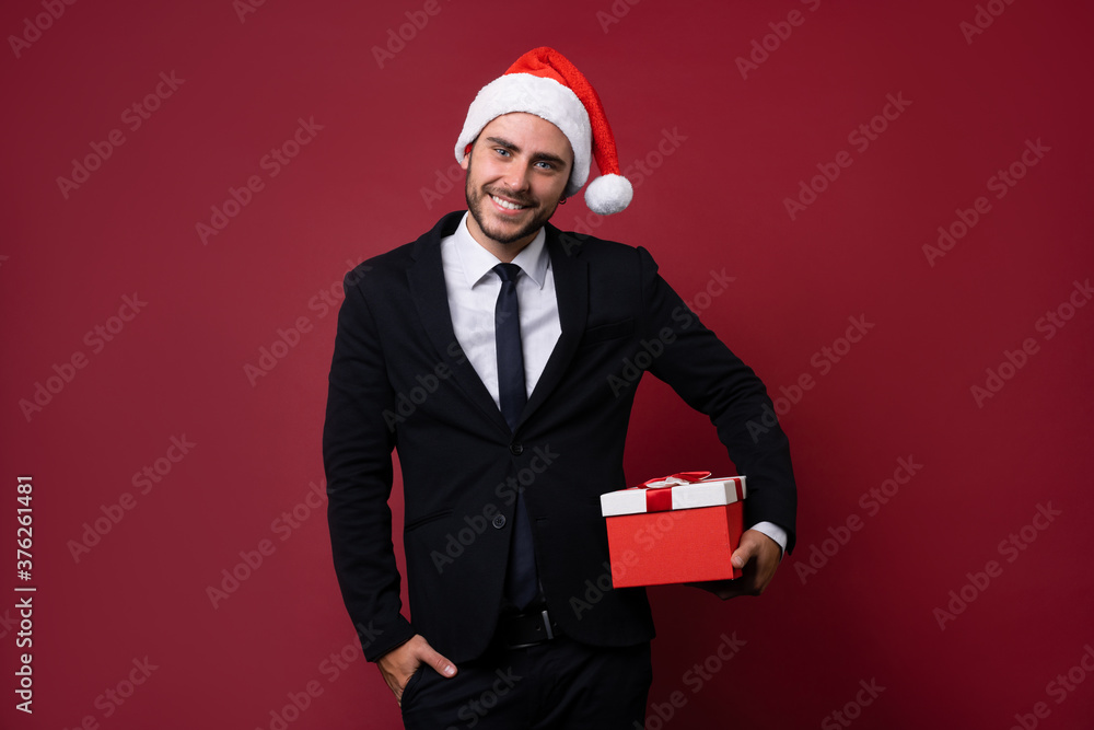 Young handsome caucasian guy in business suit and Santa hats stands on red background in studio and teeth smilie Holding red gift box in hand. Young handsome caucasian guy in business suit and Santa hats stands on red background in studio and teeth smilie Holding red gift box in hand.
