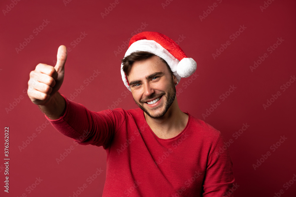 Young handsome caucasian guy in a red sweater and Santa hats stands on red background in studio and showing thumbs up. Young handsome caucasian guy in a red sweater and Santa hats stands on red background in studio and showing thumbs up.