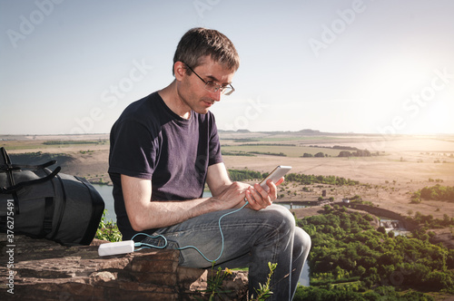 Man on a hike uses smartphone while charging from the power bank on the rock at dawn. Healthy lifestyle and communication.