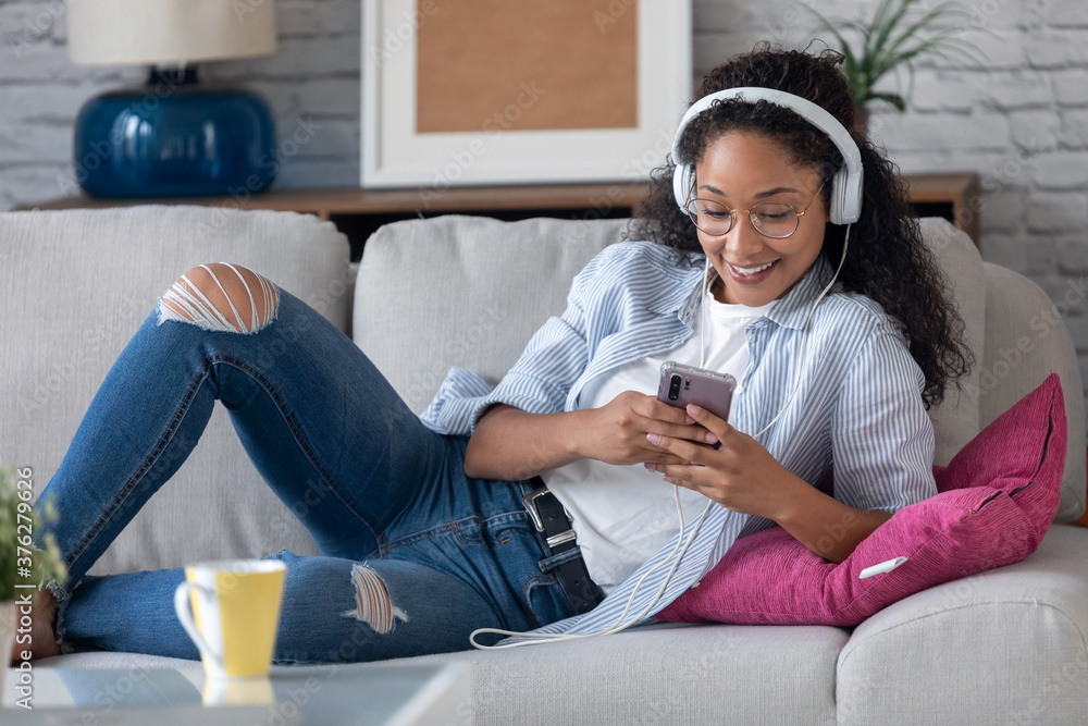 Obraz premium Beautiful young woman listening to music with headphones while using her smartphone sitting on sofa at home.