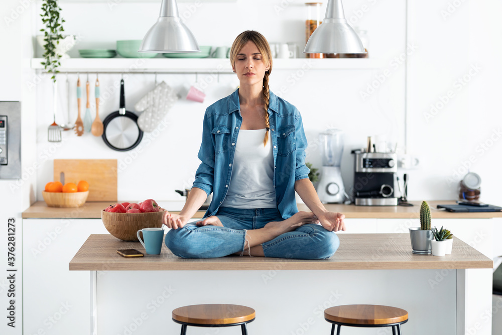 Pretty young woman in lotus position sitting on the table while relaxing in the kitchen at home. Pretty young woman in lotus position sitting on the table while relaxing in the kitchen at home.