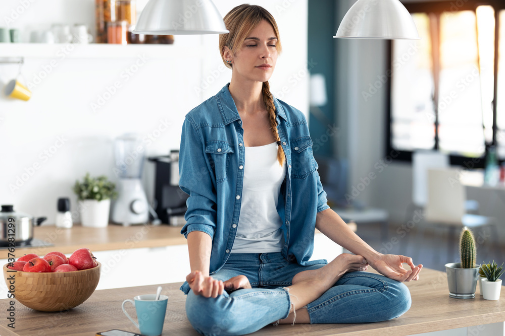 Pretty young woman in lotus position sitting on the table while relaxing in the kitchen at home. Pretty young woman in lotus position sitting on the table while relaxing in the kitchen at home.