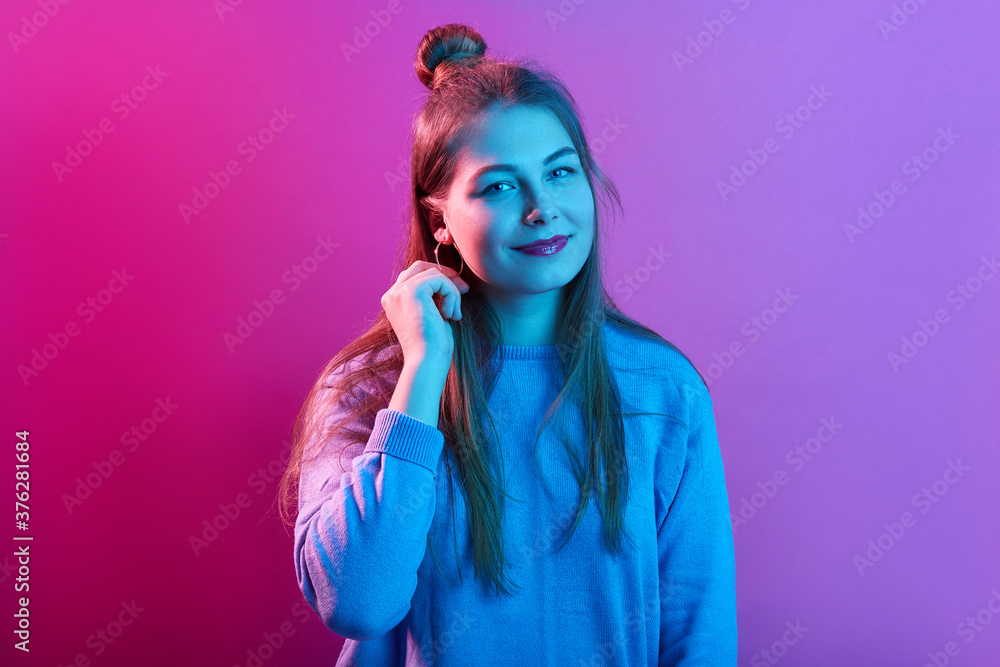 Fototapeta premium Pleasant looking young woman wears casual sweater, has hair bun, looks with calm facial expressions, poses against pink neon background, lady keeps hand near ear.