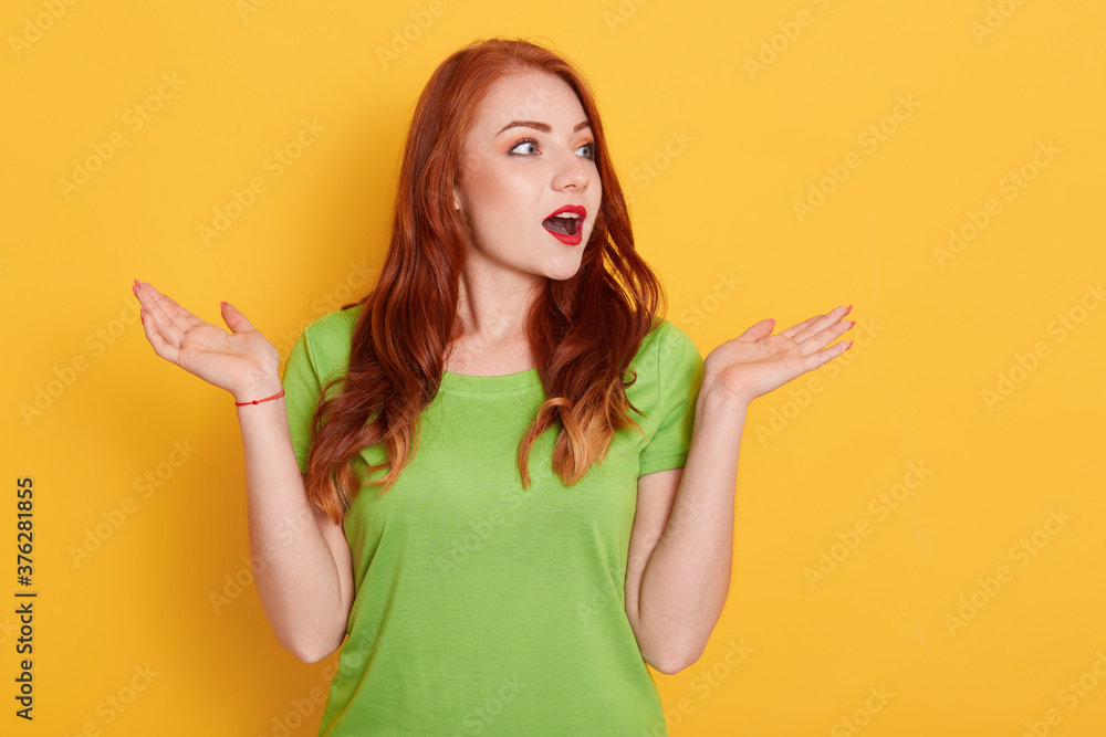 Close up portrait of beautiful red haired woman spreading hands and looking aside, standing isolated over yellow background, astonished female with widely opened mouth. Close up portrait of beautiful red haired woman spreading hands and looking aside, standing isolated over yellow background, astonished female with widely opened mouth.