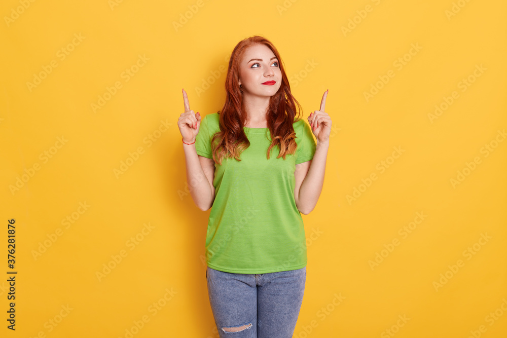 Photo of attractive dark haired young European woman pointing with index fingers up at copy space, poses against yellow background, looking aside. Photo of attractive dark haired young European woman pointing with index fingers up at copy space, poses against yellow background, looking aside.