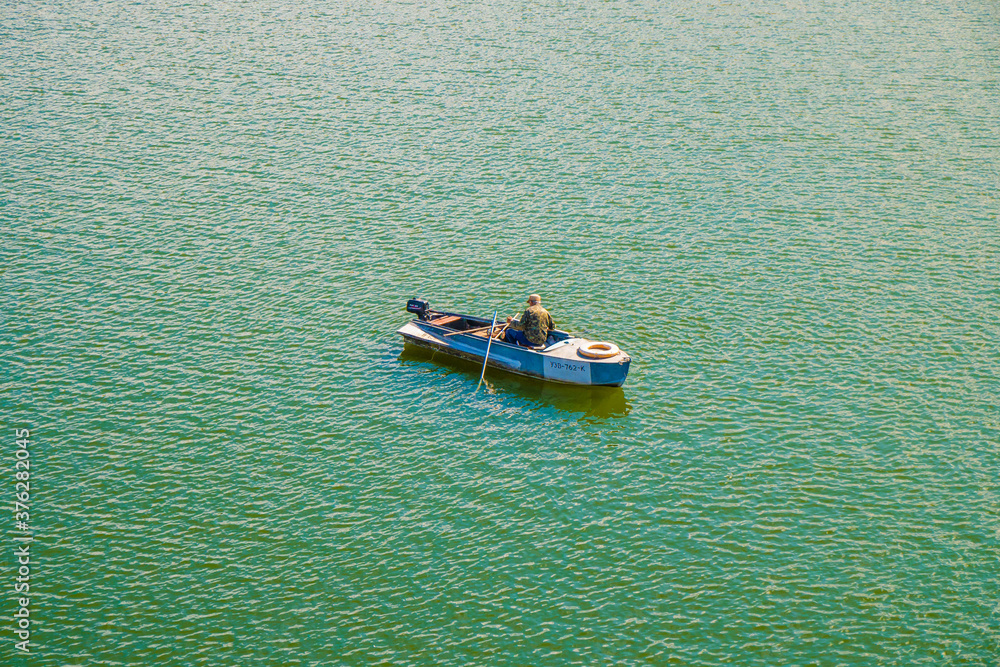 A lone fisherman in military uniform on a boat among the green water. View from above