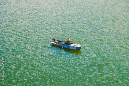 A lone fisherman in military uniform on a boat among the green water. View from above