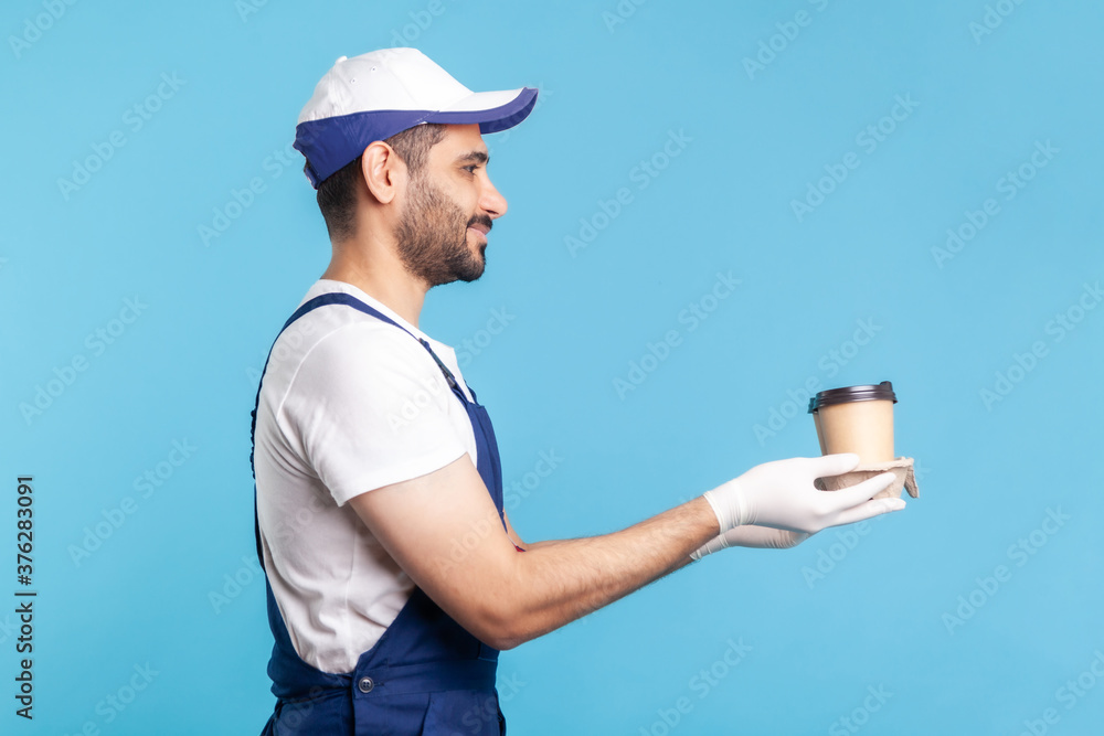 Delivery service. Side view, friendly happy courier man in overalls offering coffee, wearing safety gloves giving drinks in disposable cups and smiling. studio shot isolated on blue background Delivery service. Side view, friendly happy courier man in overalls offering coffee, wearing safety gloves giving drinks in disposable cups and smiling. studio shot isolated on blue background