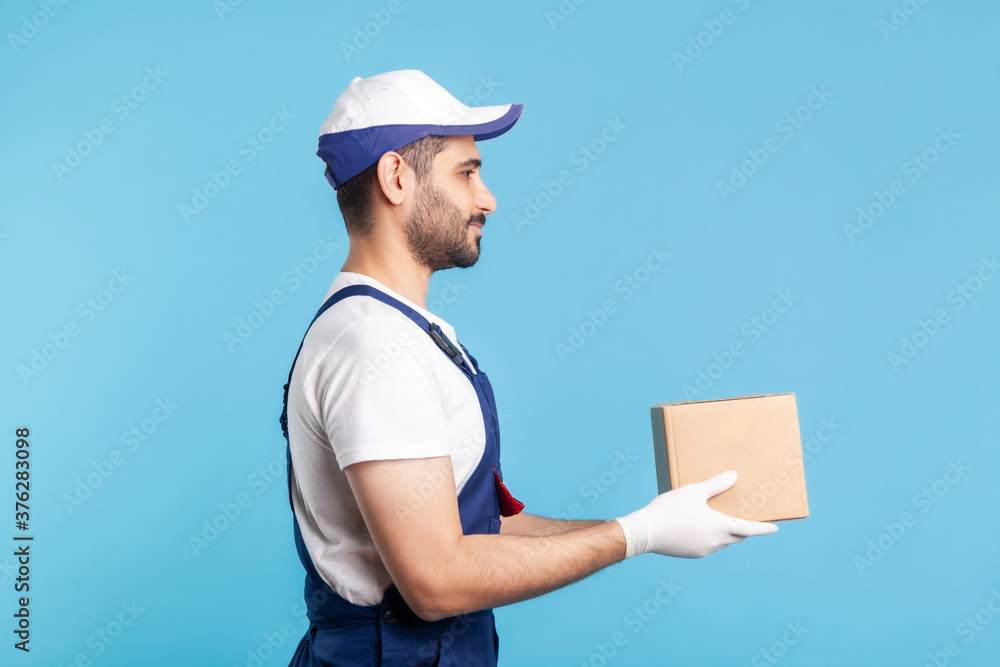 Delivery and cargo transportation. Side view, professional loader man in workwear holding cardboard box. Courier carrying parcel. Online express shipping, relocation moving services. studio shot Delivery and cargo transportation. Side view, professional loader man in workwear holding cardboard box. Courier carrying parcel. Online express shipping, relocation moving services. studio shot