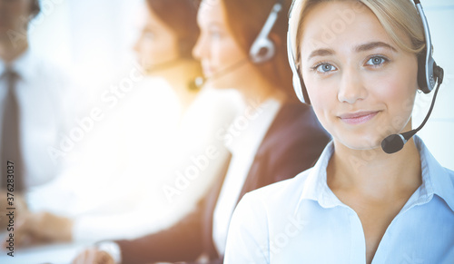 Cheerful smiling business woman with headphones consulting clients. Group of diverse phone operators at work in sunny office.Call center and business people concept