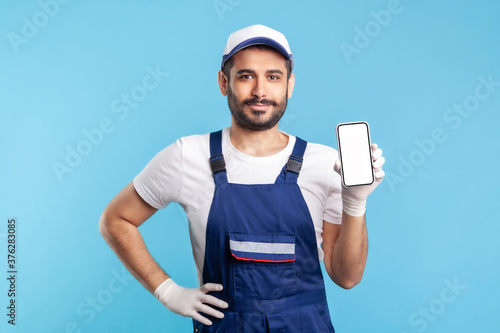 Worker in overalls and gloves holding mobile phone with blank display, device mock up for online delivery order app, house repair maintenance services. indoor studio shot isolated on blue background